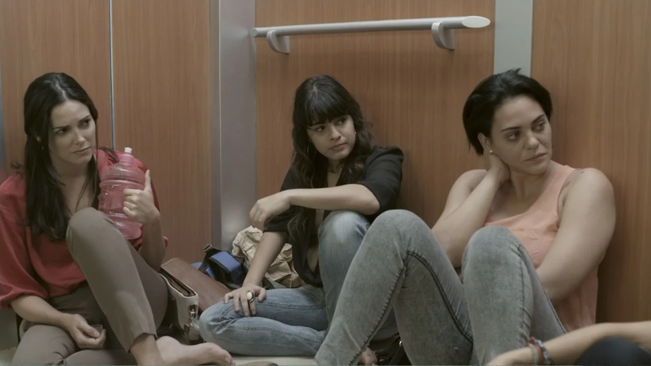 Locas y atrapadas (2014) movie still: medium — Three women are sitting on the floor inside a wood-paneled elevator car. They are positioned against…