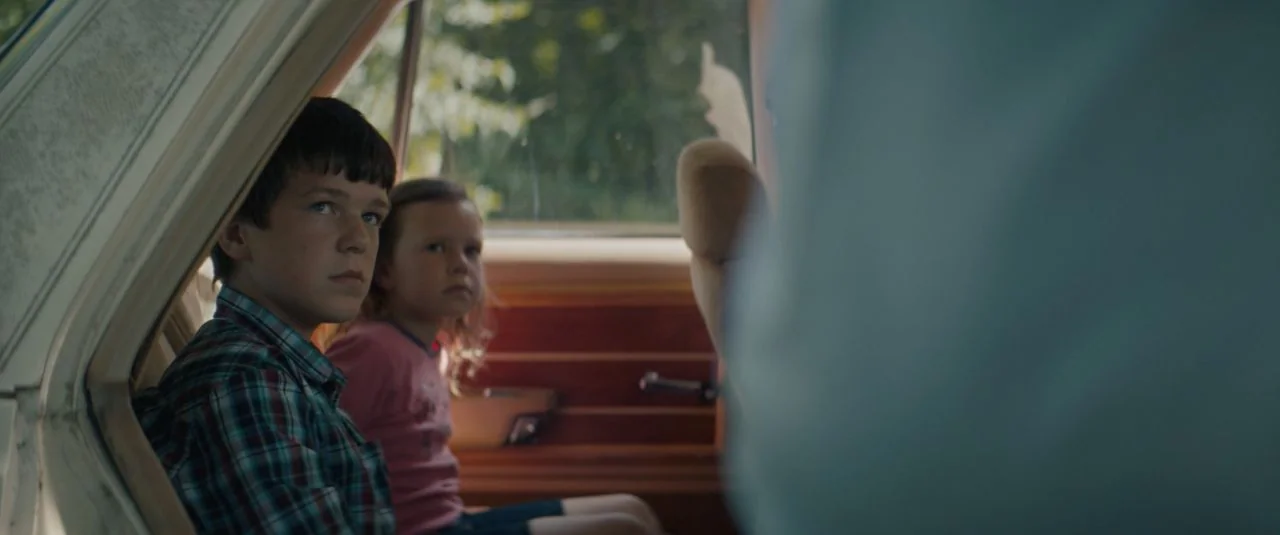 Becky (2020) movie still: medium — The interior of a moving car is visible from the backseat during the daytime. A young boy and a girl are seated…