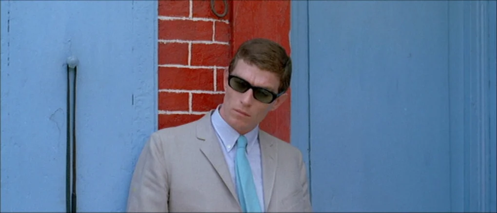 Made in U.S.A. (1966) movie still: medium — A man leans against a wall featuring red bricks framed by two blue doors. He stands squarely in front of…