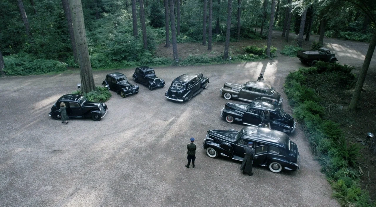The Death of Stalin (2017) movie still: wide — A gravel-surfaced clearing surrounded by tall pine trees in a European forest. Several vintage 1940s…