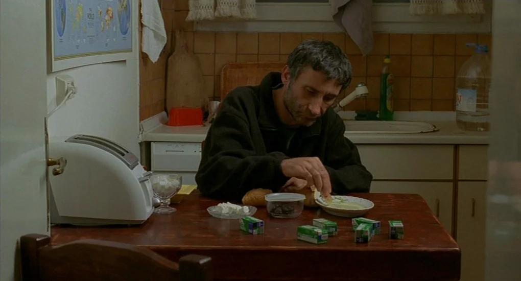 Uzak (2002) movie still: medium — A modest home area with brown tiled walls and a worn wooden table. A middle-aged man sits alone and eats a simple…