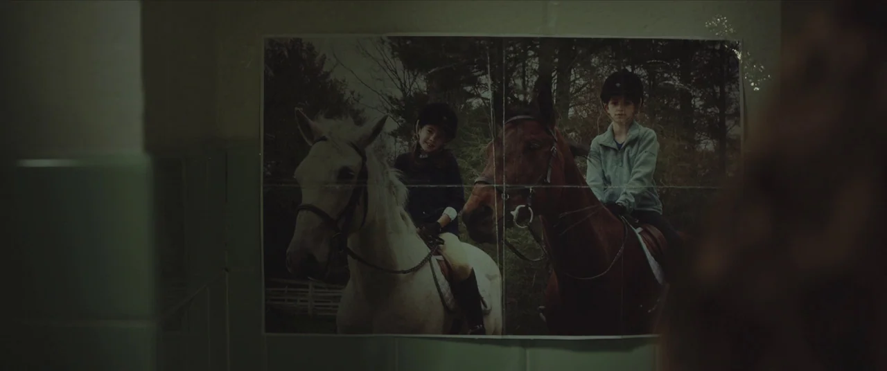 Thoroughbreds (2017) movie still: closeup — A faded photograph of two children on horseback is taped to a light-colored tiled wall. The paper is…