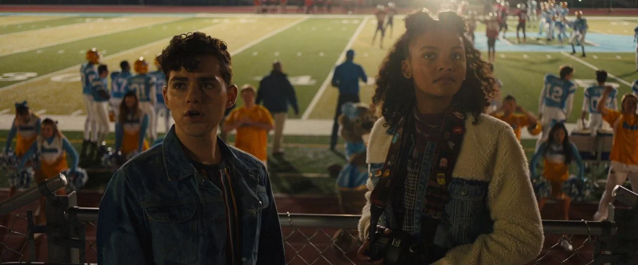 Freaky (2020) movie still: medium — A high school football stadium at night with athletes practicing on the field. Two teenagers stand behind a…