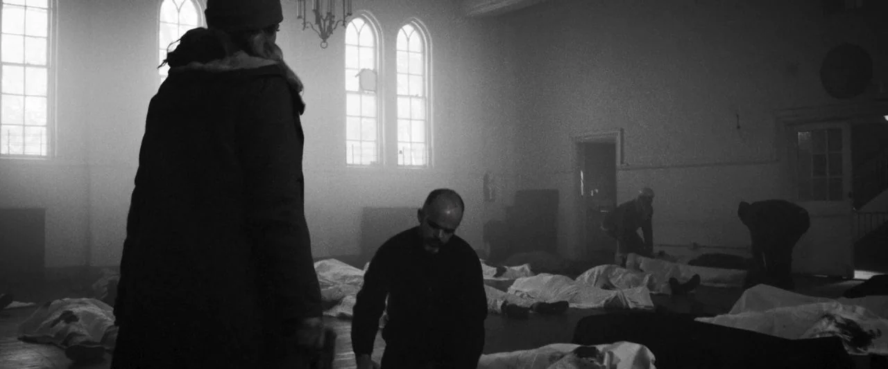 Bushwick (2017) movie still: medium — A dimly lit room with rows of people lying on the floor covered in white sheets. A man kneels in the center of…