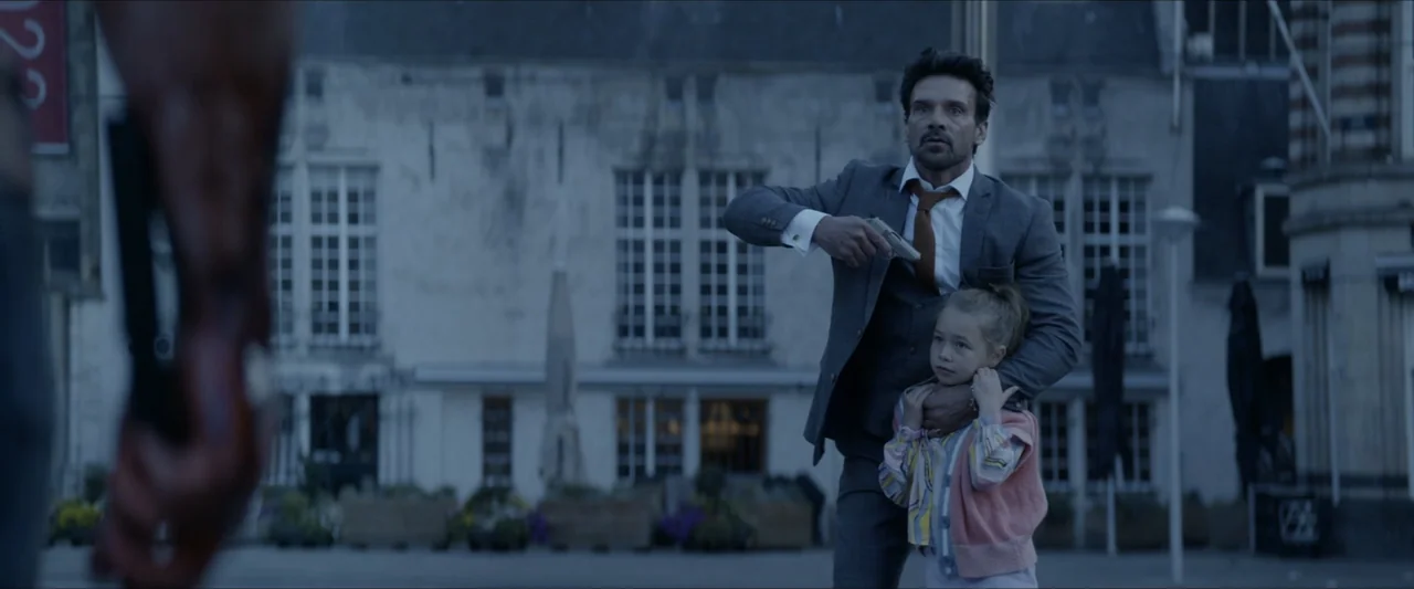 Black Lotus (2023) movie still: medium — A stone-walled public square with large windows provides the backdrop for a confrontation. A man in a suit…