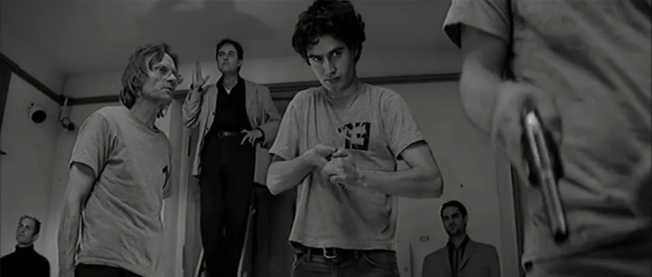 13 Tzameti (2006) movie still: medium — A sparse room with white walls and a few men standing inside. A young man loads a handgun in the center…