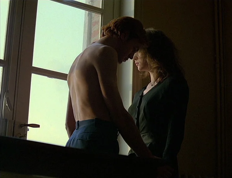First Name: Carmen (1983) movie still: medium — A dimly lit room with a large window frame letting in bright daylight. A young man and a woman stand…