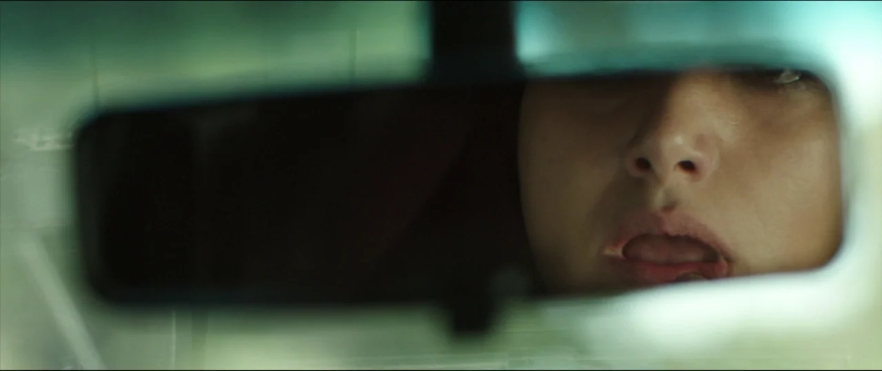 Under The Shadow (2016) movie still: closeup — The interior of a vehicle seen through a small rectangular rearview mirror frame. A person applies…