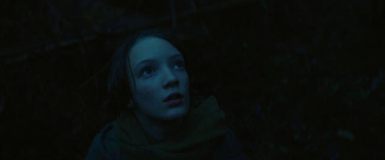 Vesper (2022) movie still: closeup — A dark forest environment at night with minimal ambient light visible in the frame. A young girl looks upward…