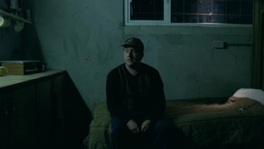 Garage (2007) movie still: medium — A dimly lit room with bare, textured walls and a simple twin bed. A man sits motionless on the edge of the…