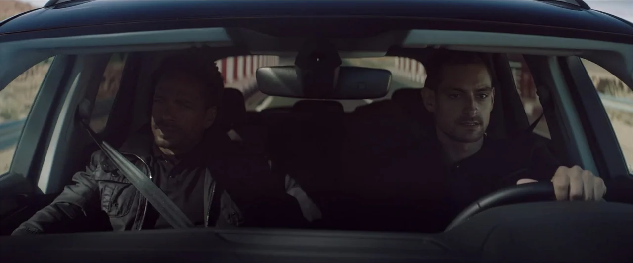 Redemption Day (2021) movie still: medium — Inside a moving car on a bridge during daylight hours. Two men are seated in the front, one driving and…
