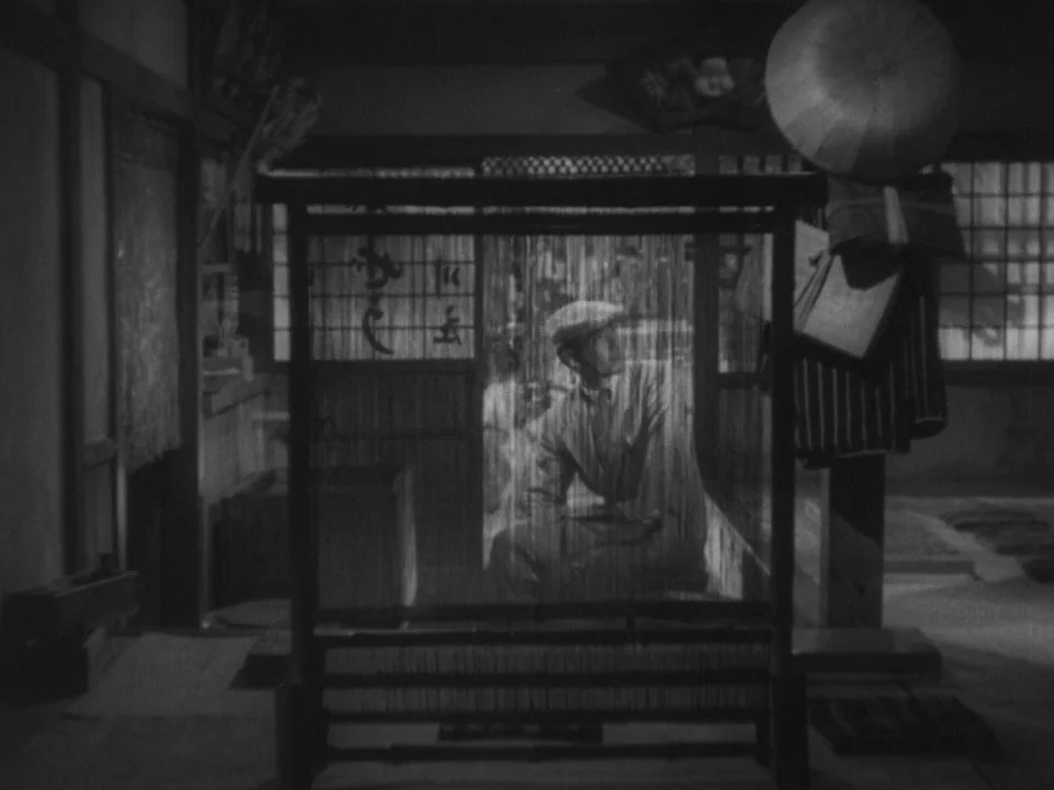 Stray Dog (1949) movie still: medium — A traditional dwelling interior is partitioned by a vertical frame loom. A man sits behind the loom,…