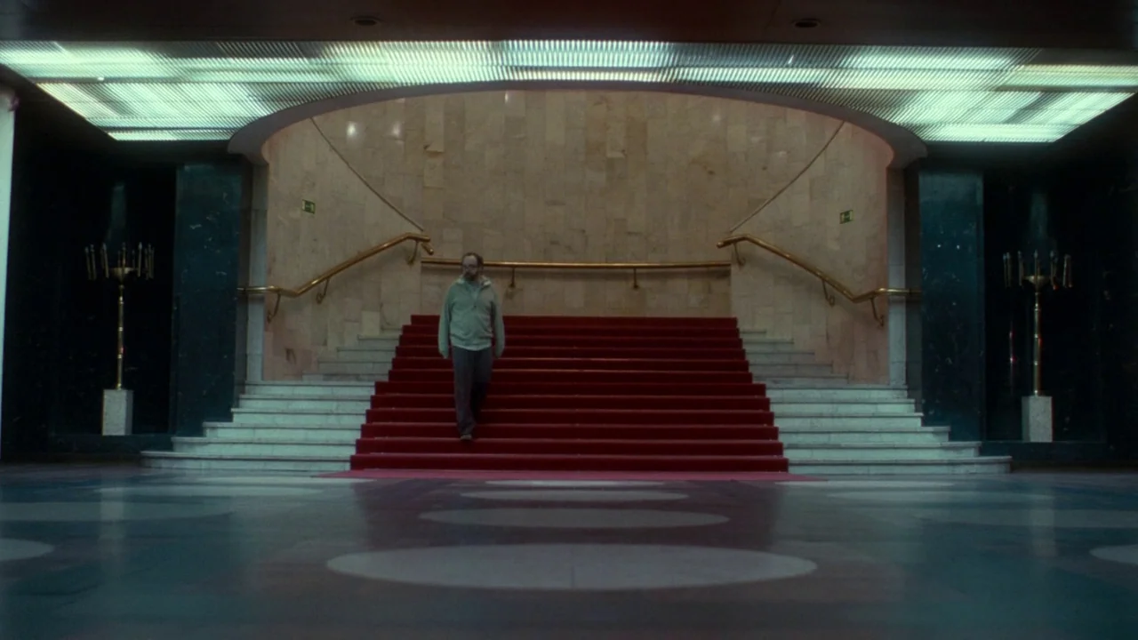 Cold Souls (2009) movie still: wide — A spacious, formal lobby features a curved stone wall and a prominent red staircase. A single man descends the…
