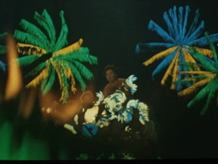 Salome (1972) movie still: medium — A nighttime parade scene with large decorative feather structures framing the subject. A performer in a detailed…
