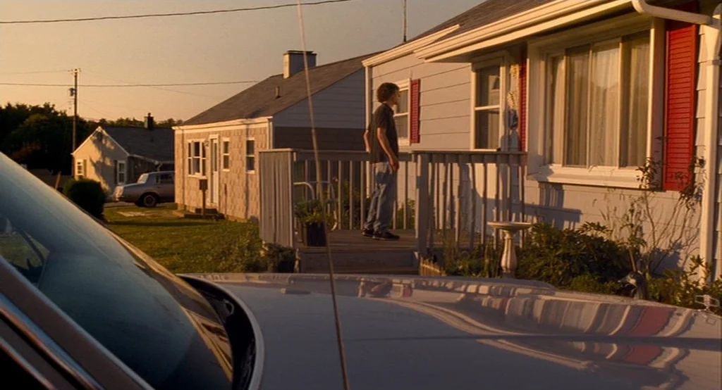 Adventureland (2009) movie still: wide — A suburban residential street with blue-sided houses and red shutters during golden hour. A young man…