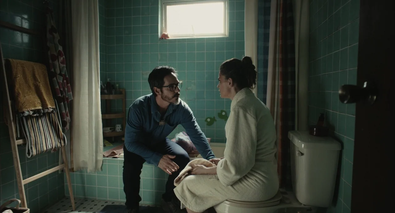 The Diary (1999) movie still: medium — A small bathroom with light teal tiled walls and a small frosted window above. A man kneels on the edge of a…