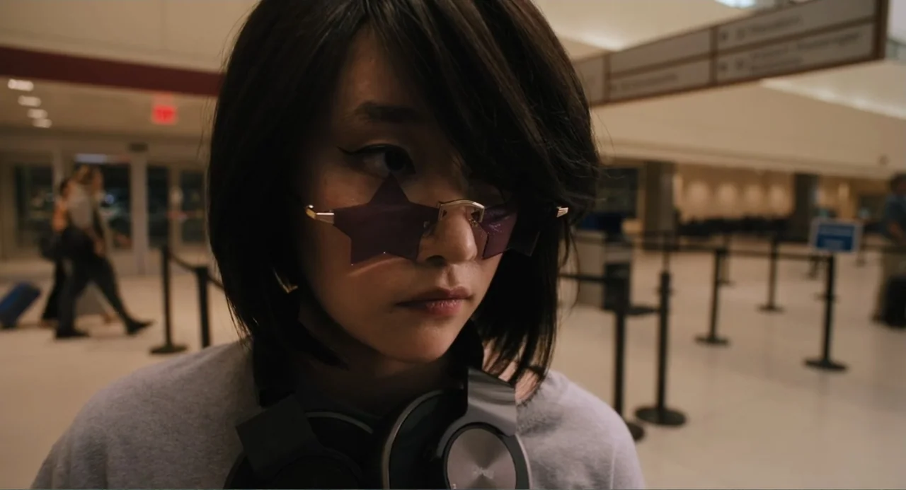 Mona Lisa and the Blood Moon (2022) movie still: closeup — A young woman stands inside a brightly lit, sterile airport terminal with queue barriers.…