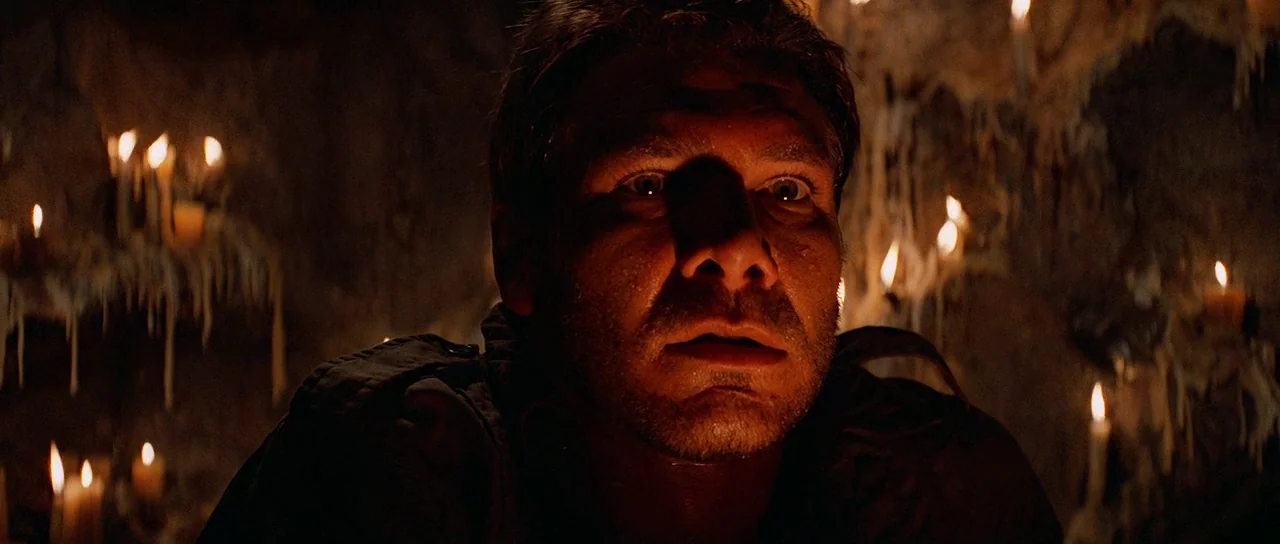 Indiana Jones and the Temple of Doom (1984) movie still: closeup — A dimly lit, rocky enclosure illuminated by multiple burning wax candles. A man…