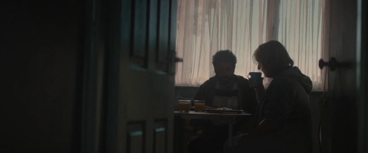 Ali & Ava (2021) movie still: medium — A dimly lit room with a man and woman at a table by a window. They eat breakfast while silhouetted against…