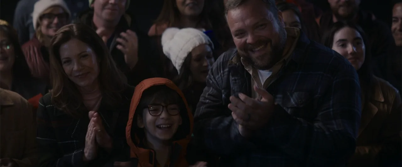 A Christmas Mystery (2022) movie still: medium — A dimly lit auditorium filled with an audience of seated people. A man, woman, and child in the…