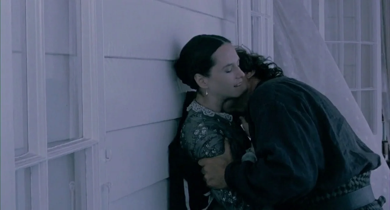 The Piano (1993) movie still: medium — A couple stands on a porch with white clapboard siding and windows behind them. They are embraced tightly…
