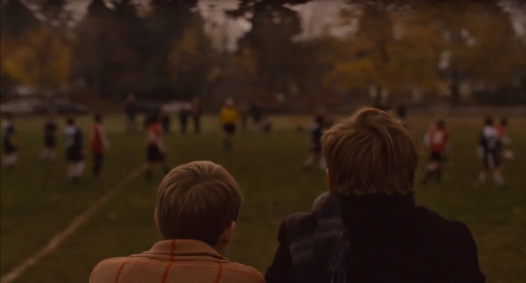 Restless (2011) movie still: medium — A grassy athletic field with players in the distance under an overcast sky. Two boys sit with their backs to…
