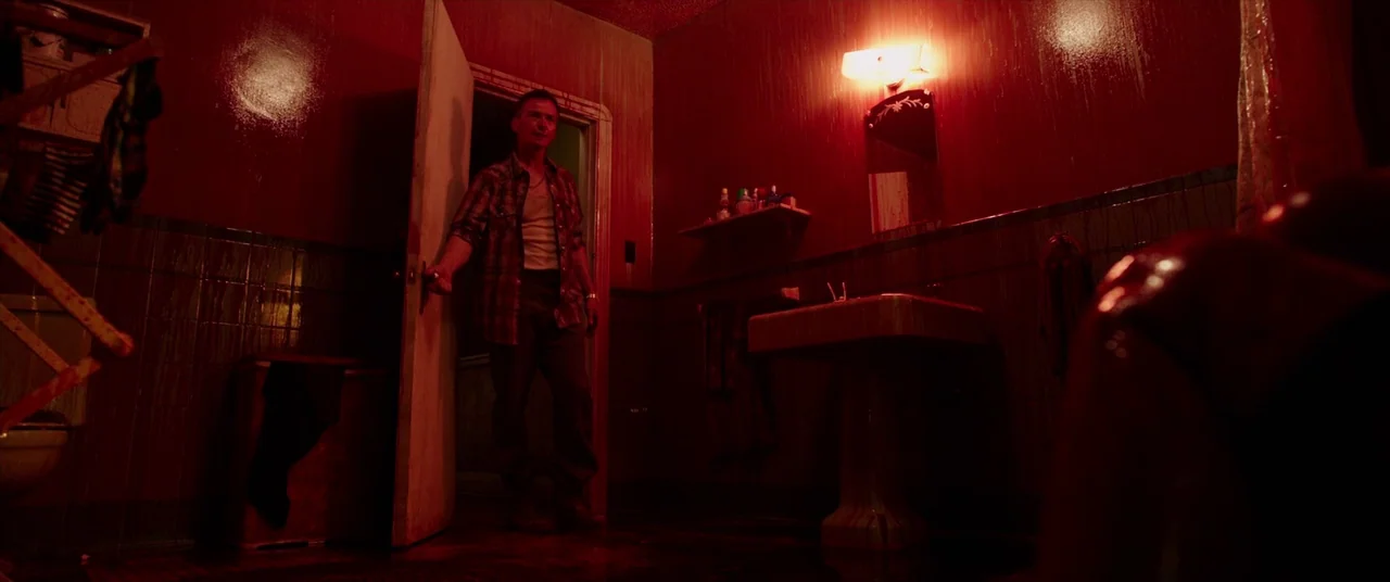 It (2017) movie still: fullbody — A dark, tile-walled washroom is illuminated by a single glowing red light fixture. A man stands at the threshold…