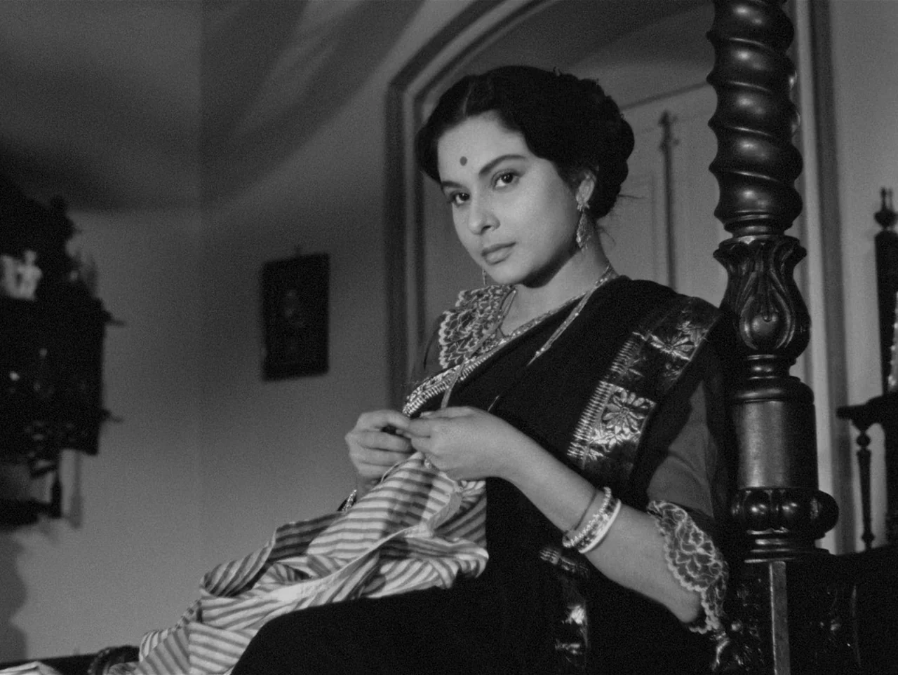 Charulata (1964) movie still: medium — An interior space features a dark ornate wooden bedpost beside a seated woman. She is sewing a piece of…