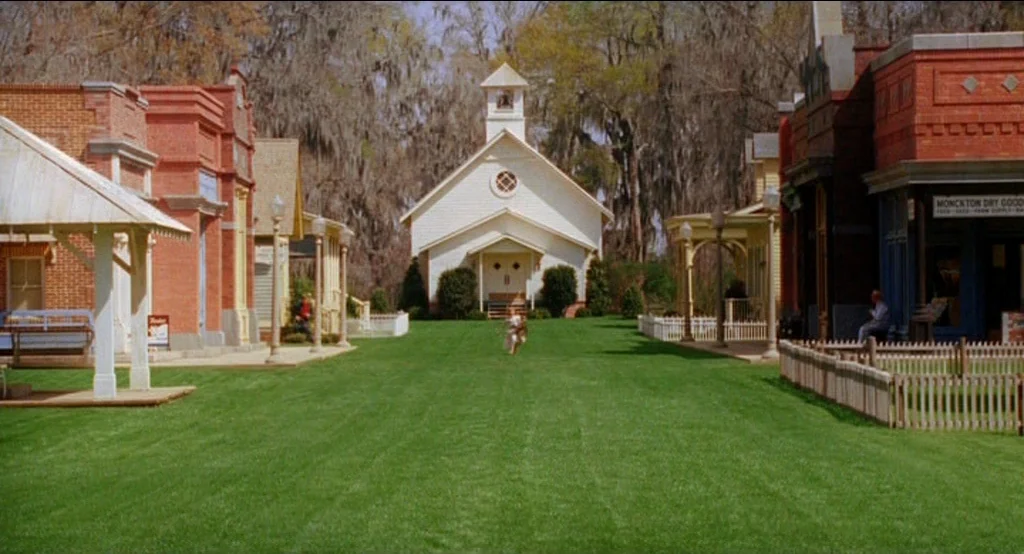 Big Fish (2003) movie still: wide — A small town square features a large lawn centered on a white church building. A young boy runs across the green…