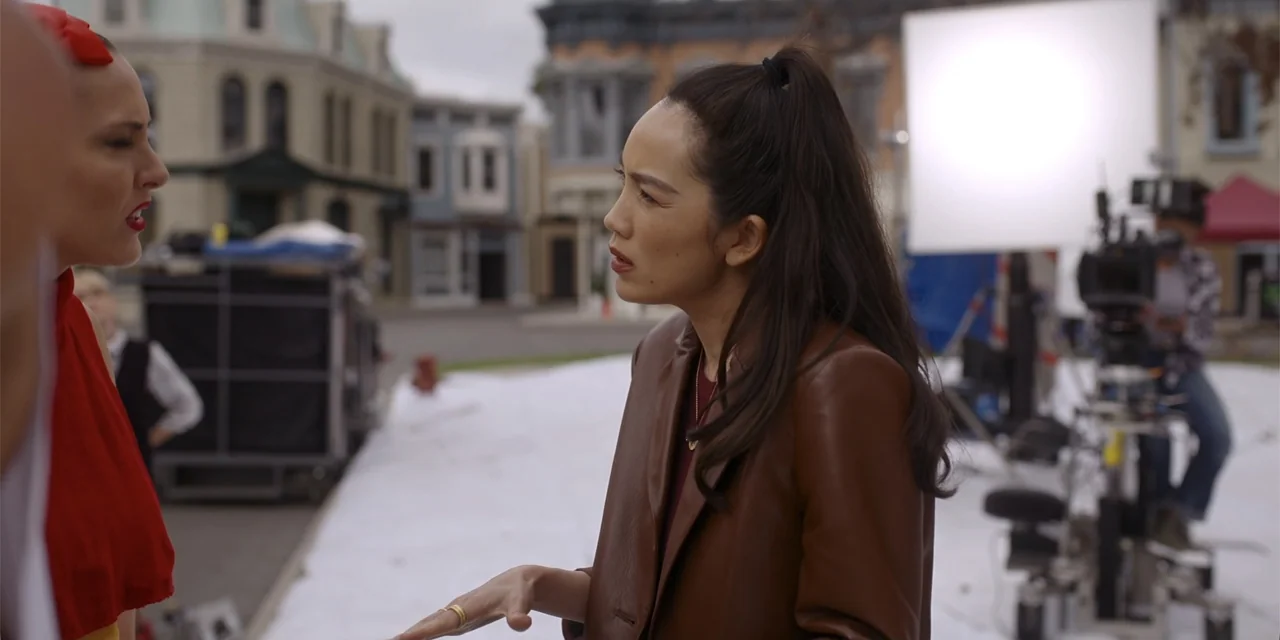 A Hollywood Christmas (2022) movie still: medium — A film production lot features background buildings and equipment visible around two women. The…
