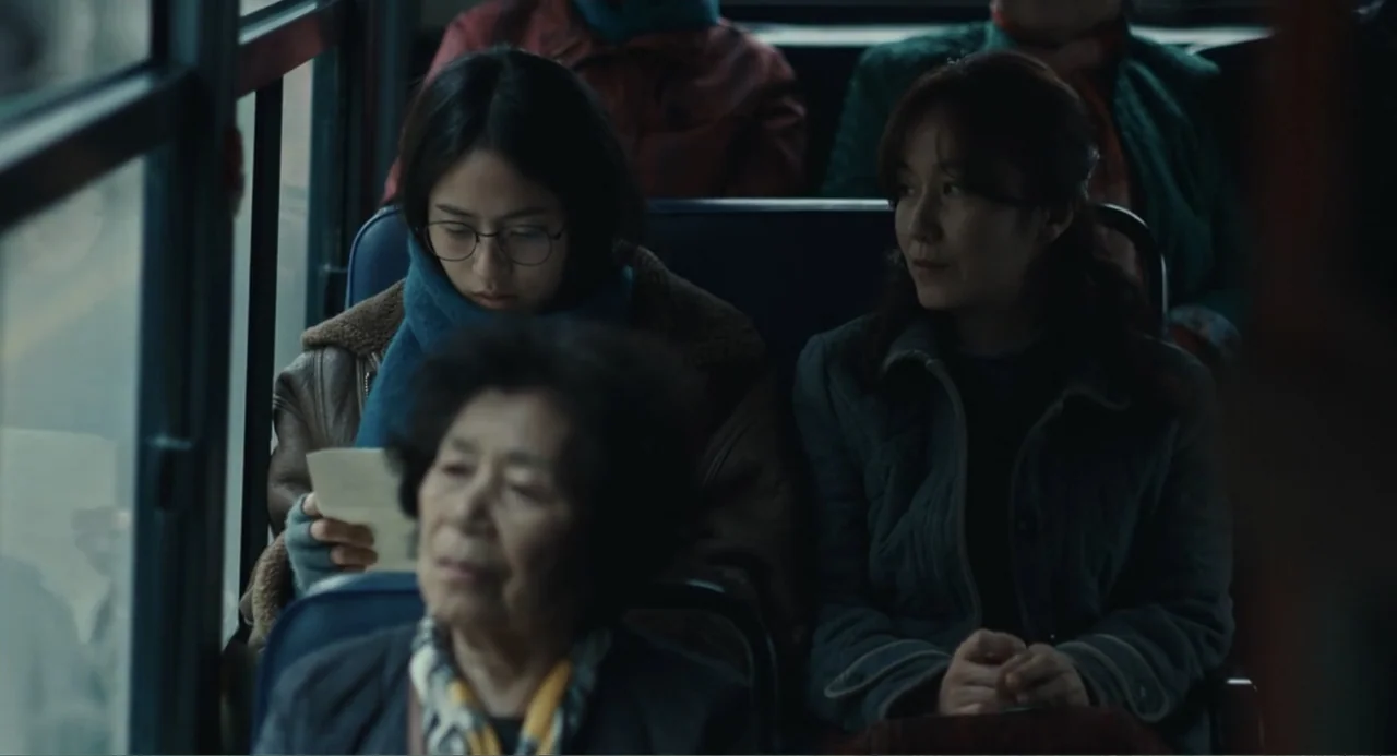 Winter in Sokcho (2025) movie still: medium — The interior of a public bus with rows of blue padded seats. Three women sit in various seats while…