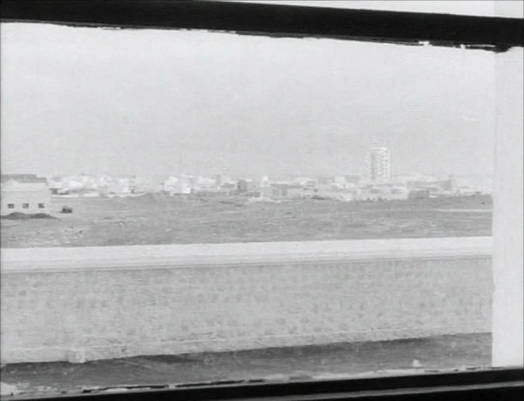 Even Dwarfs Started Small (1970) movie still: establishing — A view through a window showing a vast, sparse city landscape under a pale sky. A long…