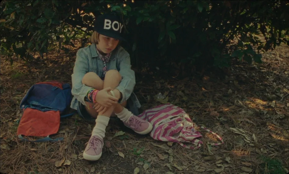 Misunderstood (2014) movie still: fullbody — A child sits under the shade of a dense tree on a leaf-covered floor. She wears a black baseball cap…