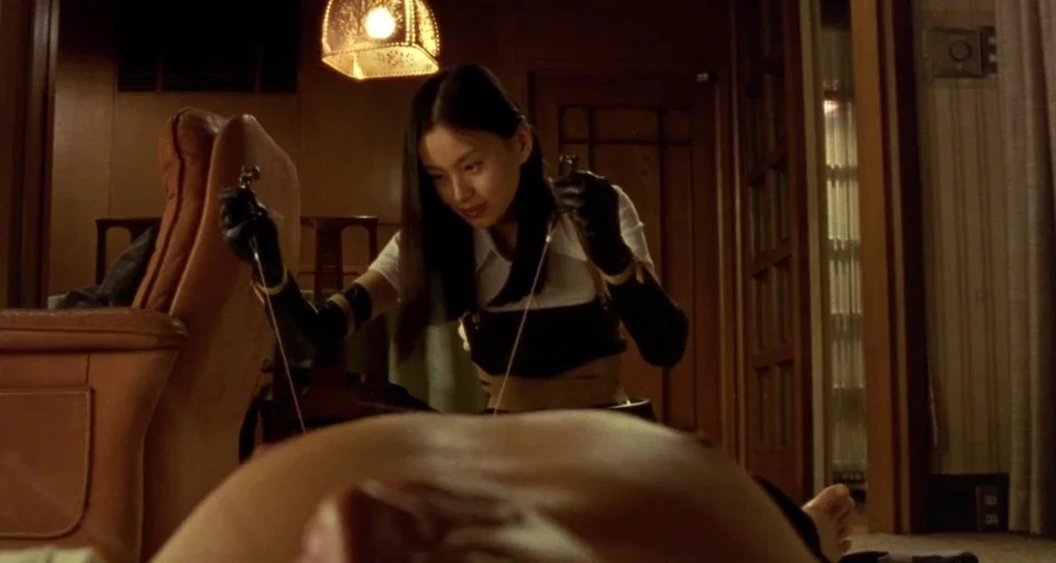 Audition (1999) movie still: medium — An indoor apartment features wooden furniture and an overhead pendant light with patterned lampshade. A young…
