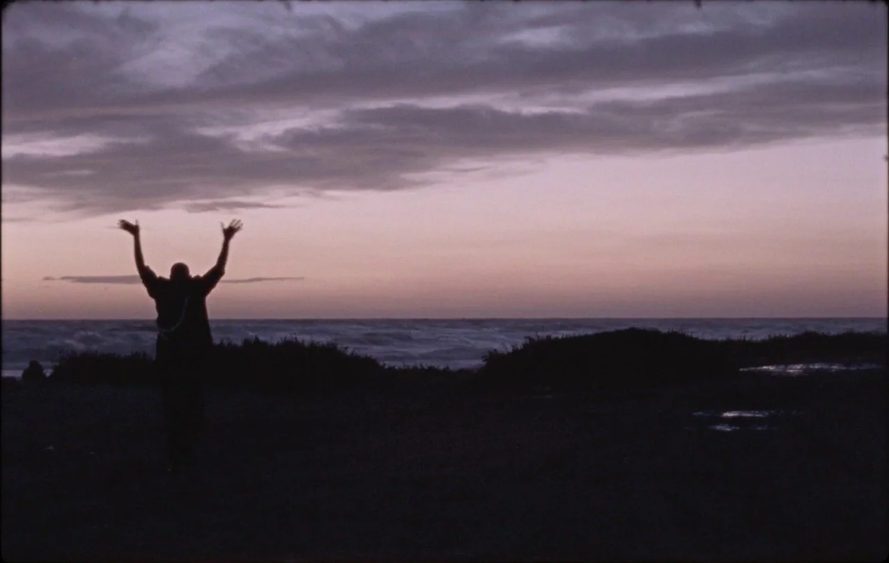 La Chimera (2023) movie still: wide — A dark coastline at dusk with rolling waves under a pastel purple cloud layer. A silhouetted person stands on…