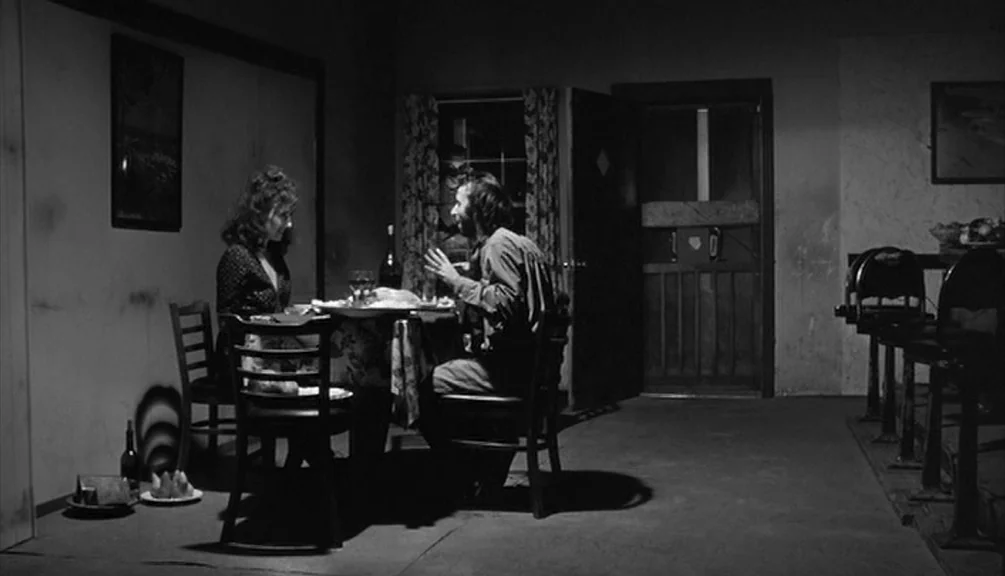 Down By Law (1986) movie still: wide — A dark, sparsely furnished room with a wooden table and chairs in the center. A man and a woman sit facing…