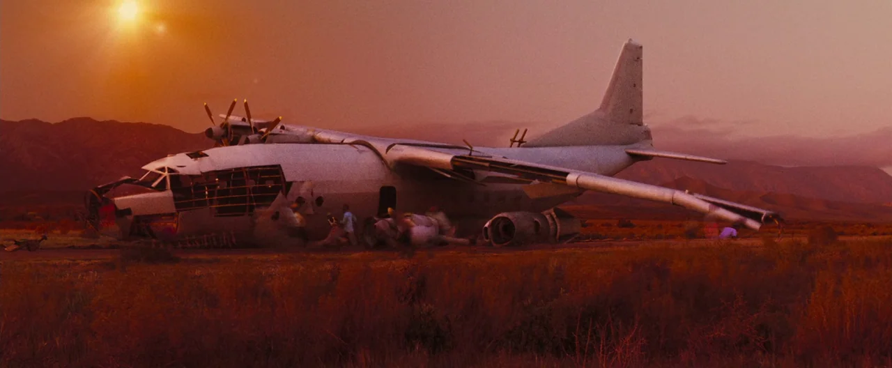 Lord of War (2005) movie still: wide — A damaged airplane sits in a dry, sparse desert at sunset. The fuselage is breached and resting on the sandy…