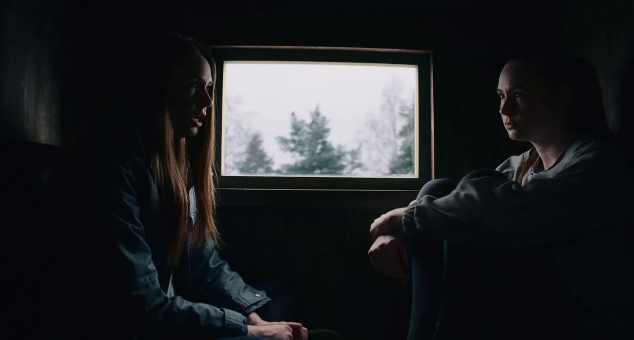 Dual (2022) movie still: medium — Two women sit inside a dark wooden structure facing a single rectangular window. The window reveals a blurry…