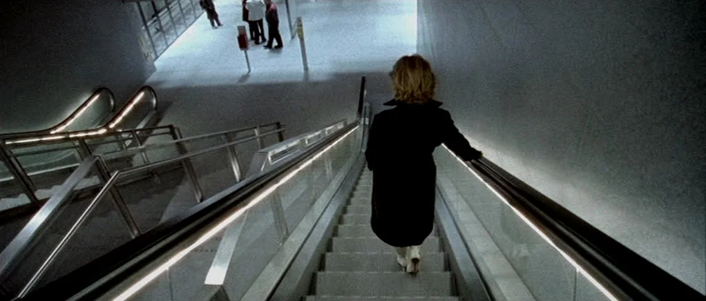 Reconstruction (2003) movie still: medium — A modern subway station interior features clean grey walls and metallic escalator railings. A person in…
