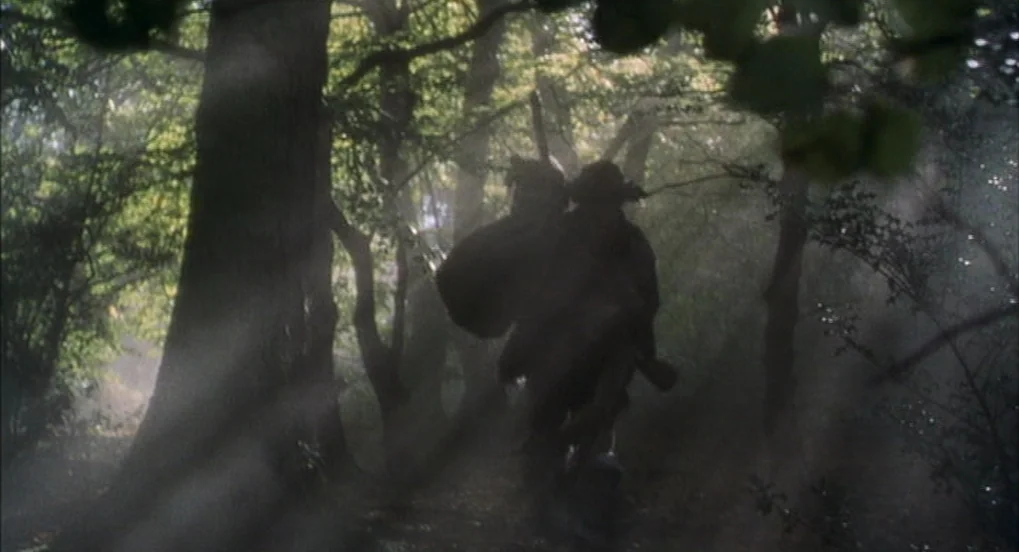 Jabberwocky (1977) movie still: fullbody — A dense woodland area is filled with thick ground fog and shafts of light. A cloaked figure walks away…
