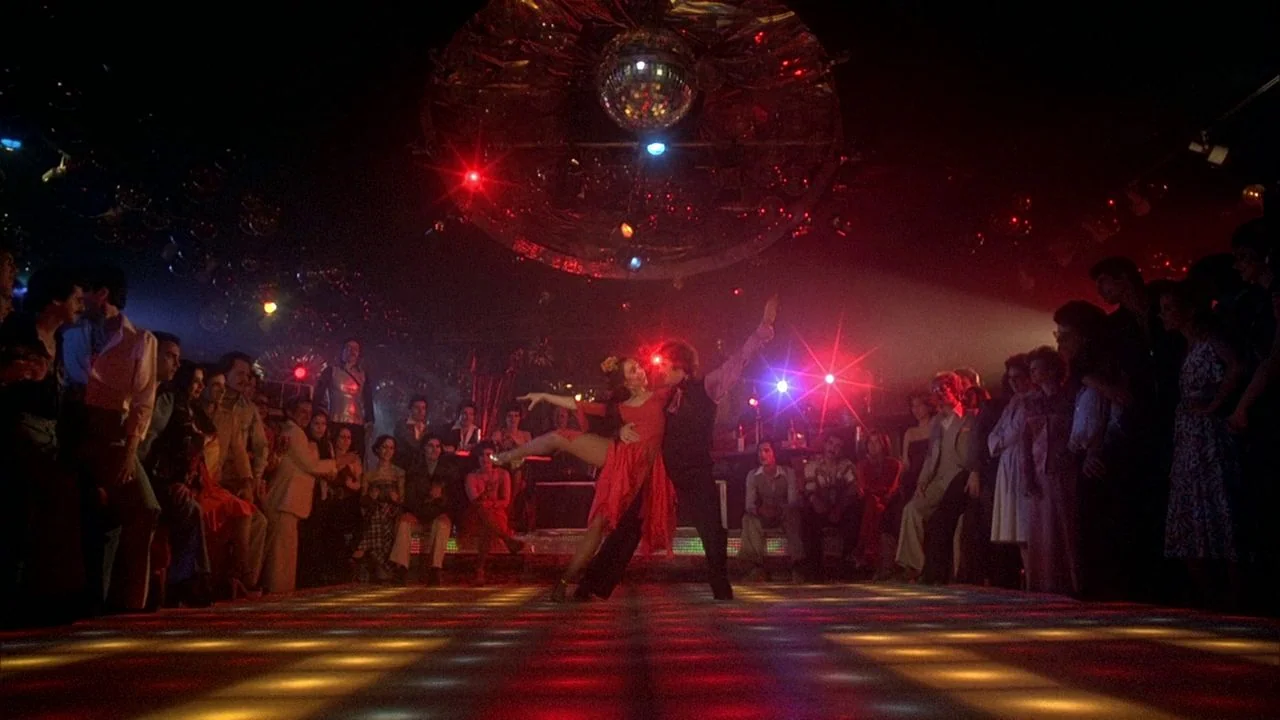 Saturday Night Fever (1977) movie still: wide — A dark 1970s nightclub interior features a large disco ball hanging from the ceiling. A couple…