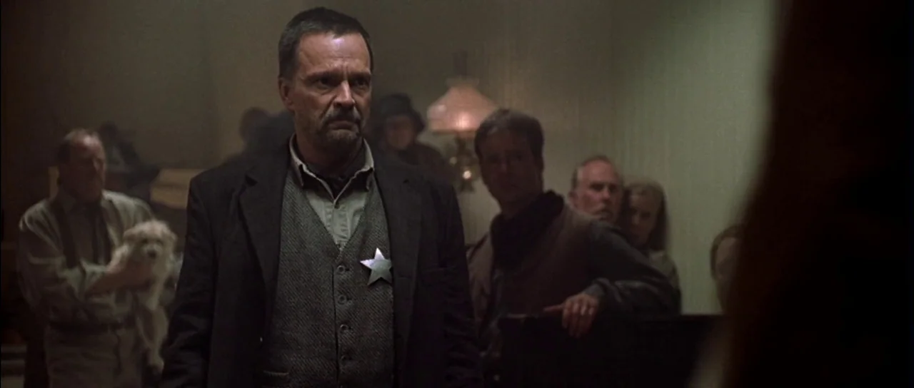 Open Range (2003) movie still: medium — A dimly lit meeting room has wooden paneling and several figures gathered in shadow. A man with a sheriff…
