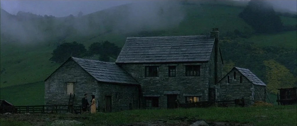 Jude (1996) movie still: wide — A stone farmhouse sits at the base of a large, mist-covered green hill. A man and a woman stand close together in…