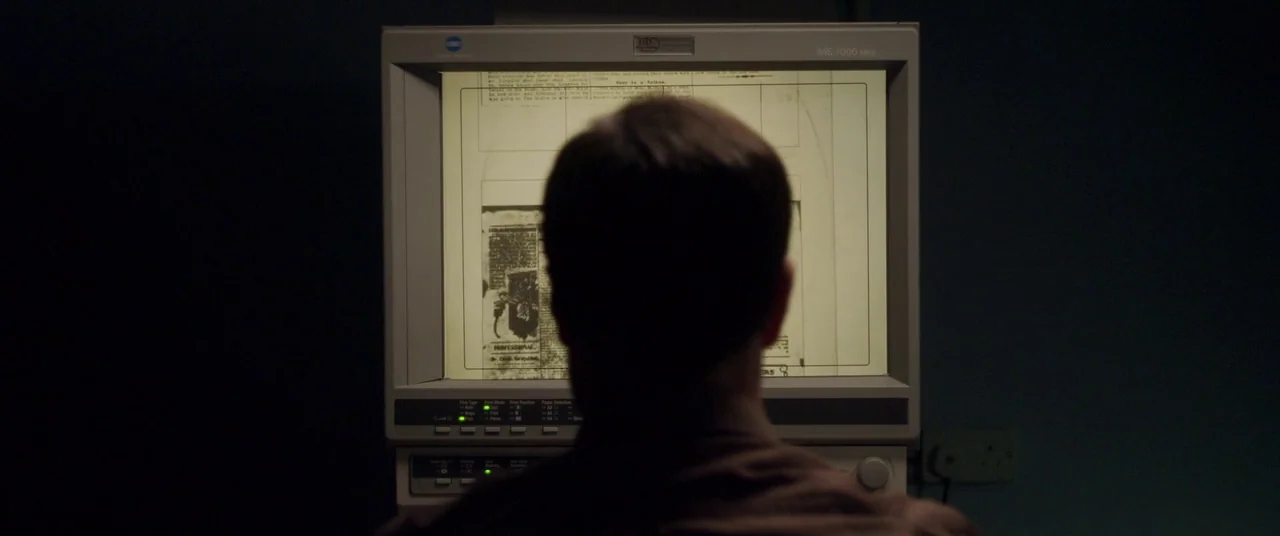The Canal (2014) movie still: medium — A dimly lit room contains a person seated before a vintage microfilm scanner device. The subject focuses…