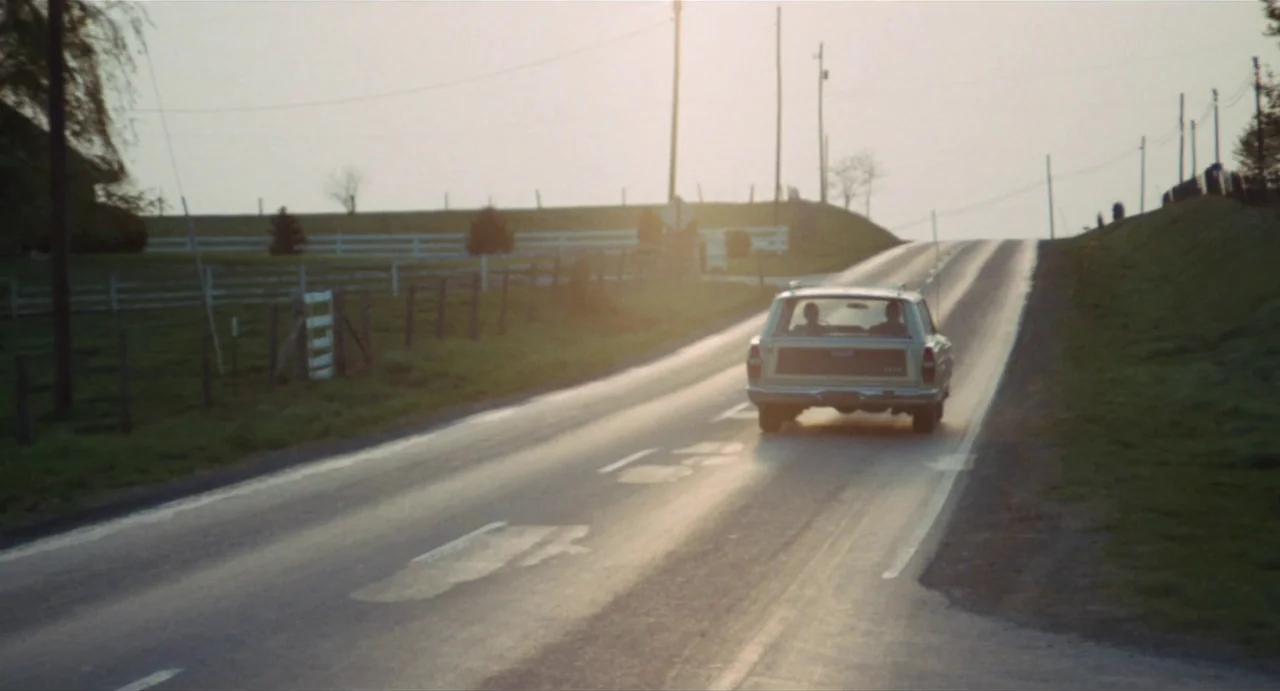The Rain People (1969) movie still: wide — A quiet two-lane country road stretches toward a distant rising slope during golden hour. A wood-paneled…