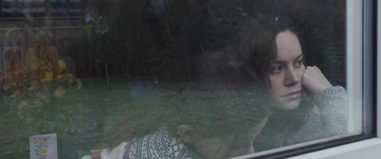 Room (2015) movie still: medium — A woman and a child gaze outside through a glass window into a yard. The window surface shows faint reflections of…