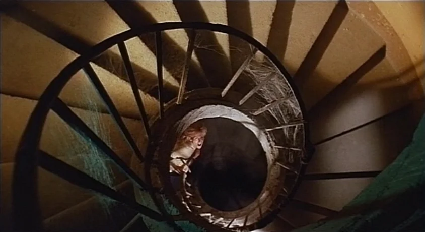 Kill, Baby Kill (1966) movie still: wide — A narrow stone spiral staircase is covered in cobwebs and cast in heavy shadow. A woman ascends the…