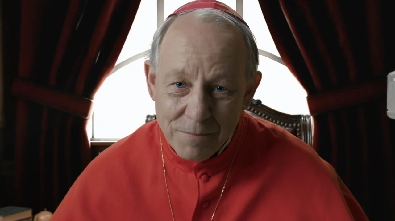Resurrected (2023) movie still: closeup — A man in red clerical robes sits in front of a window with dark drapes. The subject faces forward against…