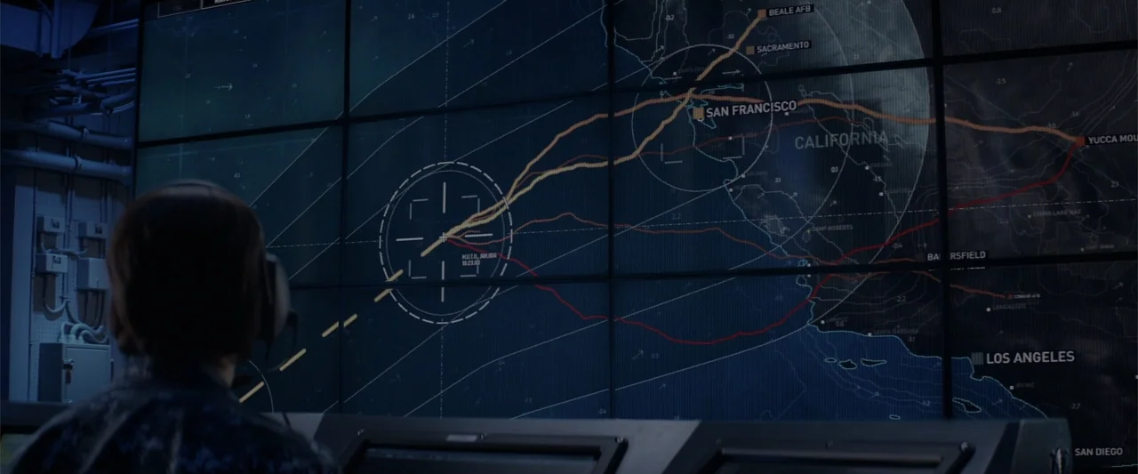 Godzilla (2014) movie still: medium — A dimly lit command center features a massive wall-mounted screen displaying tactical map data. A woman in a…