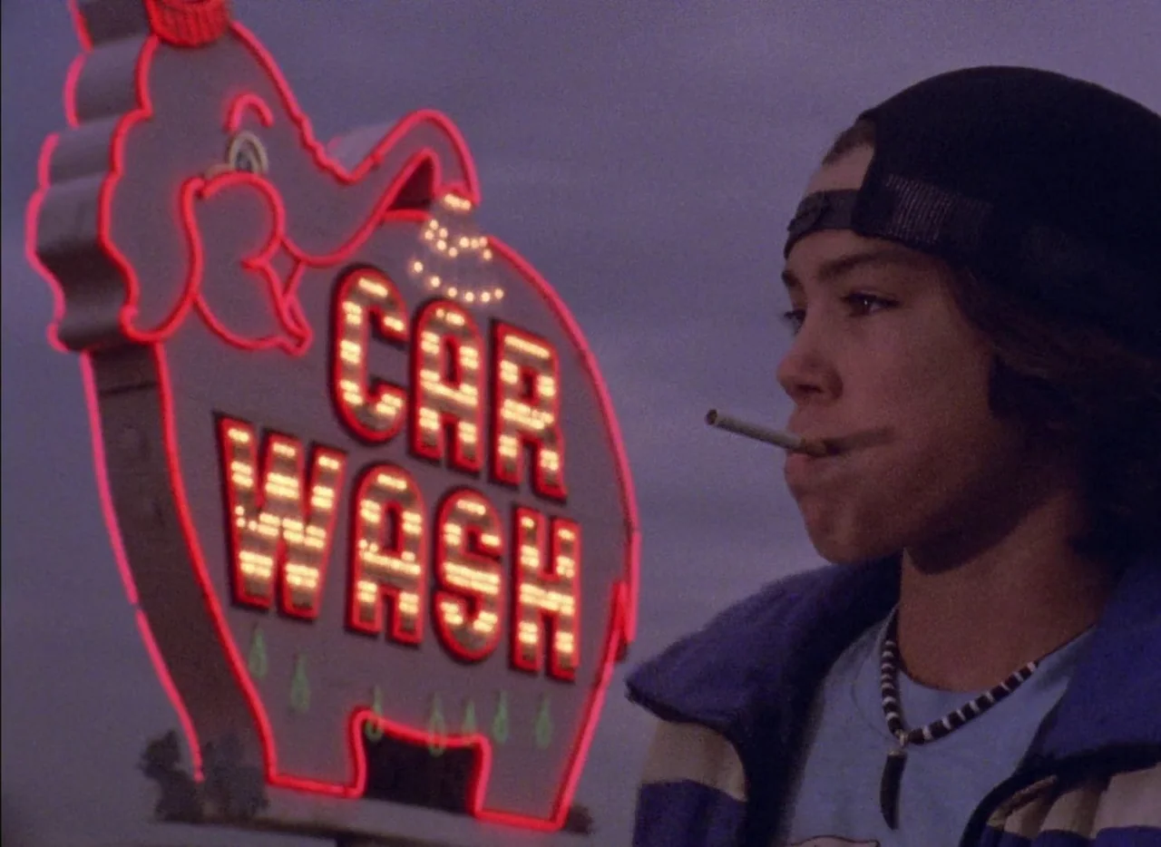 Streetwise (1984) movie still: medium — A teenager stands outdoors at dusk beside an illuminated pink elephant neon sign. The bright red sign…